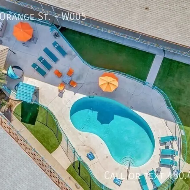 Aerial view of a swimming pool surrounded by lounge chairs and orange umbrellas, along with a grassy area nearby.