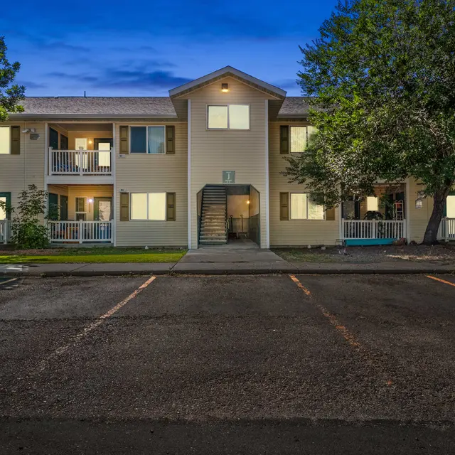 Evening view of Courtyards at Sheridan Apartments a two-story apartment building in Sheridan, WY with exterior lights illuminating balconies, entryways, and pathways. The building creates a warm, welcoming atmosphere as dusk settles, providing a cozy residential setting with ample parking.