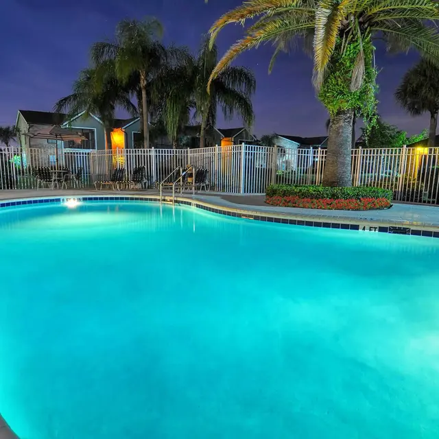 Night time view of a swimming pool surrounded by a white fence, trees, and chairs.