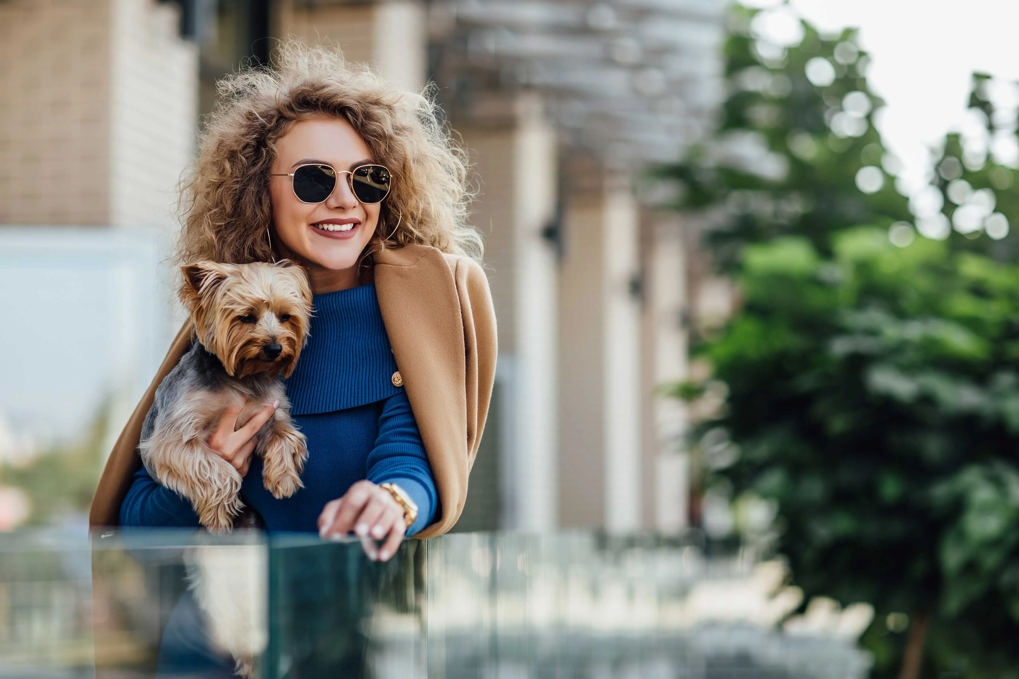 Stylish Woman with Dog Outdoors A smiling woman with curly hair wearing sunglasses holds a small dog in her arms while standing next to a glass railing in an outdoor setting.