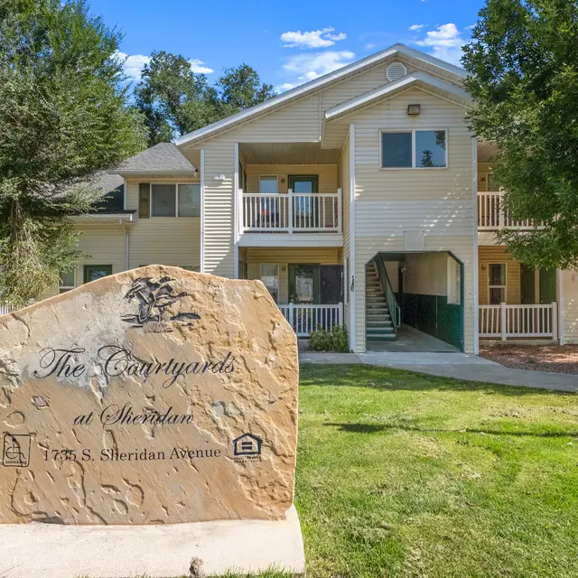 Exterior view of The Courtyards at Sheridan apartment complex, featuring a welcoming entrance with balconies and surrounded by mature trees. A large engraved stone displays the property name and address, adding a distinctive touch to the landscaped setting.