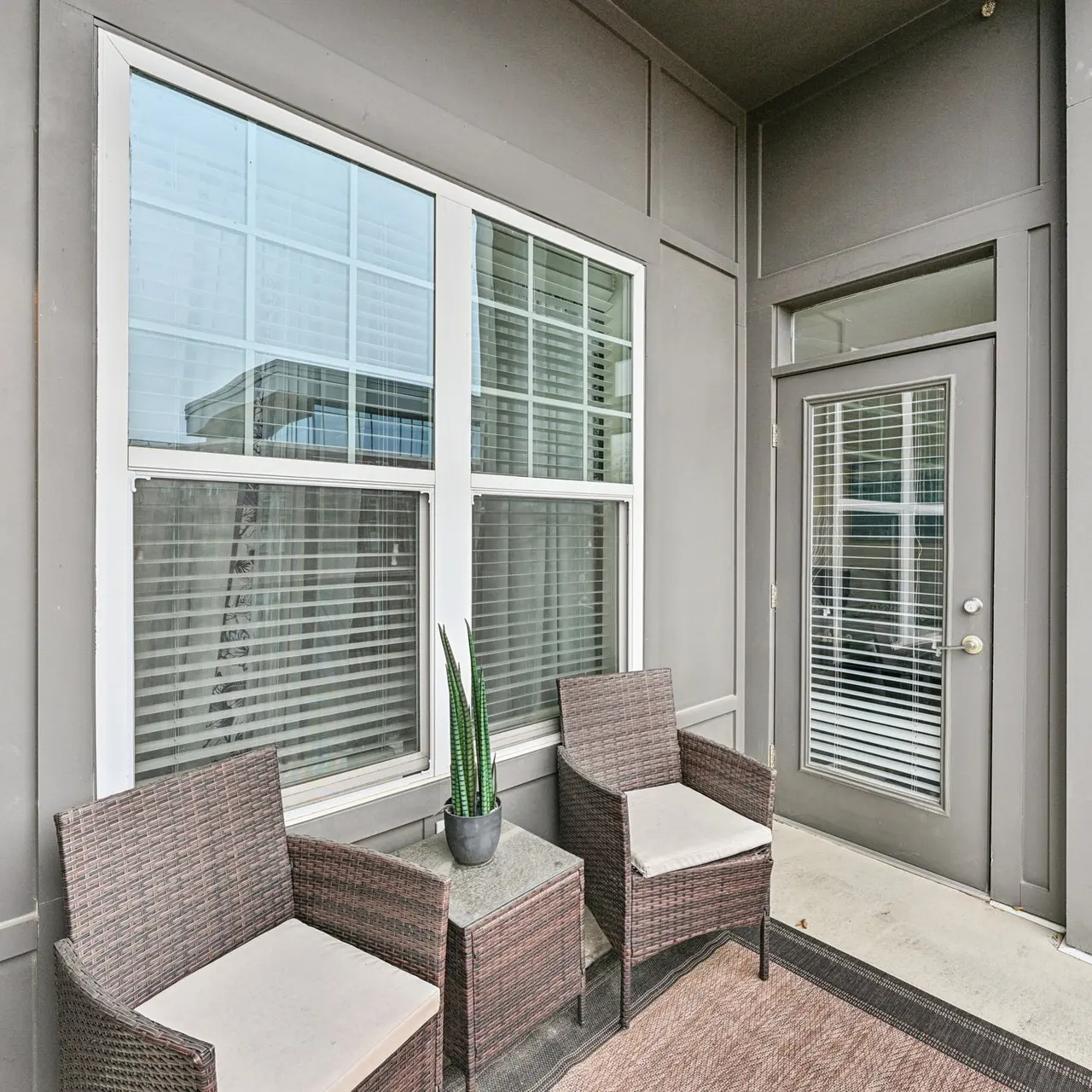 A modern balcony featuring two wicker chairs and a small potted plant next to a door leading inside.