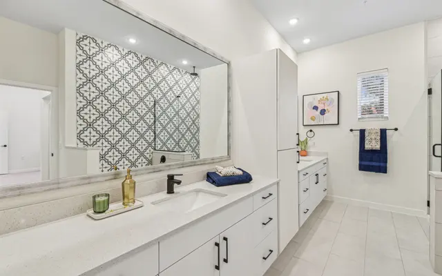 A spacious modern bathroom featuring a long white countertop with a sink, a decorative mirror, and minimalist cabinetry. The wall behind the sink is adorned with a patterned tile, and there are decorative elements like a candle and framed artwork.