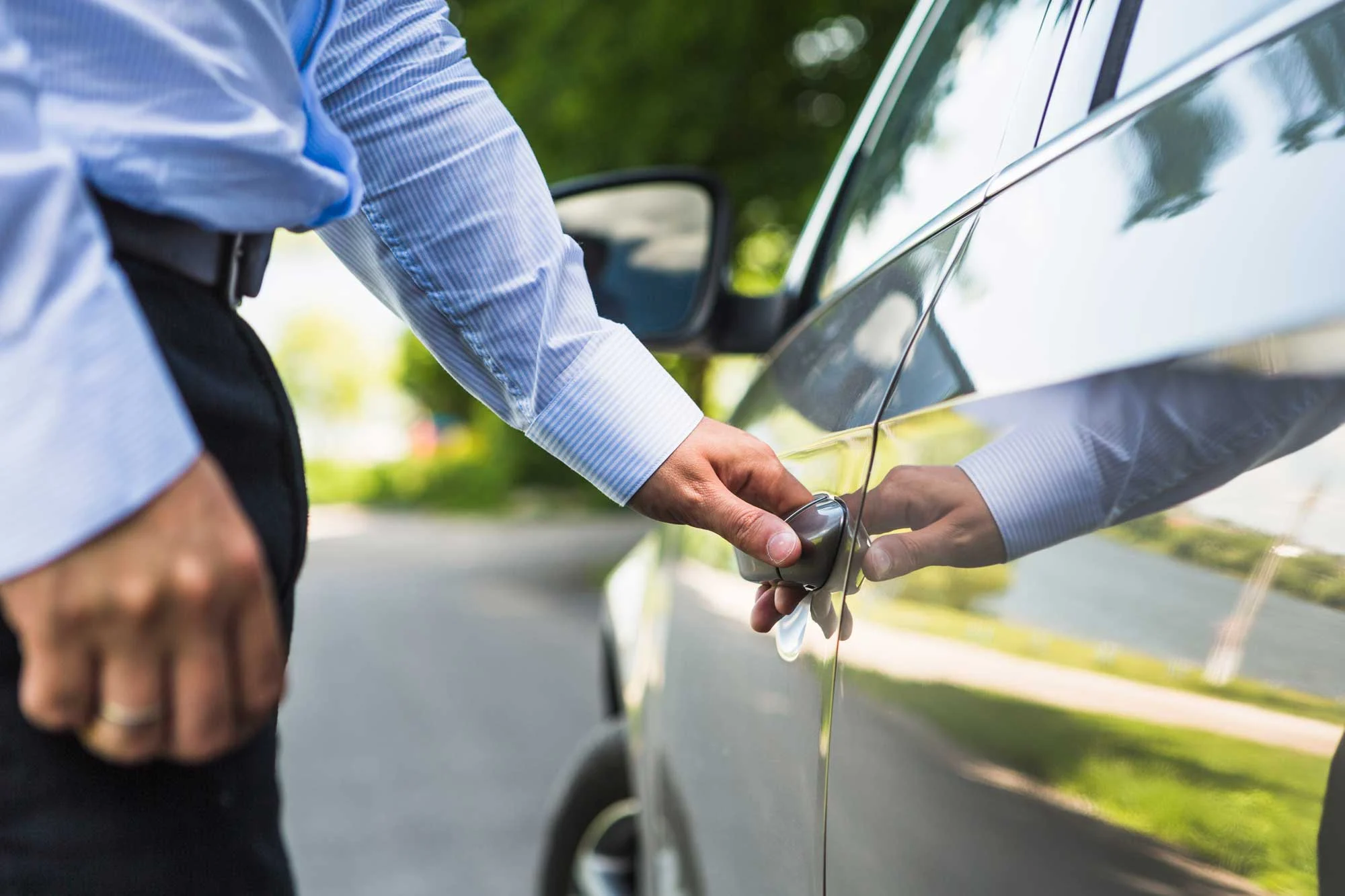 Man Opening Car Door A close-up of a man in a light blue shirt reaching to open a car door, with a blurred background of trees and a road.