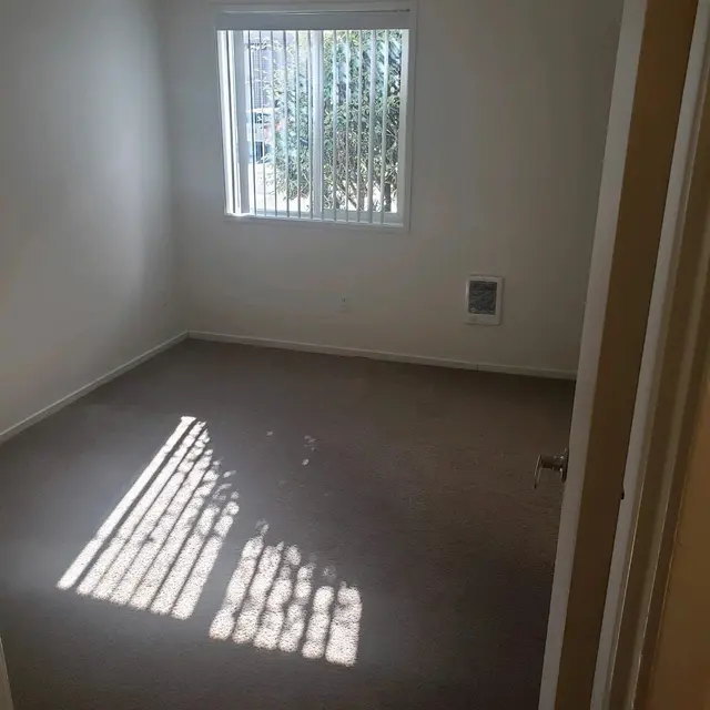 Empty Room with Natural Light A vacant room with beige carpet and a large window allowing natural light to enter. The window has vertical blinds and casts striped shadows on the floor. There is an air vent on one wall and no furniture in the room.
