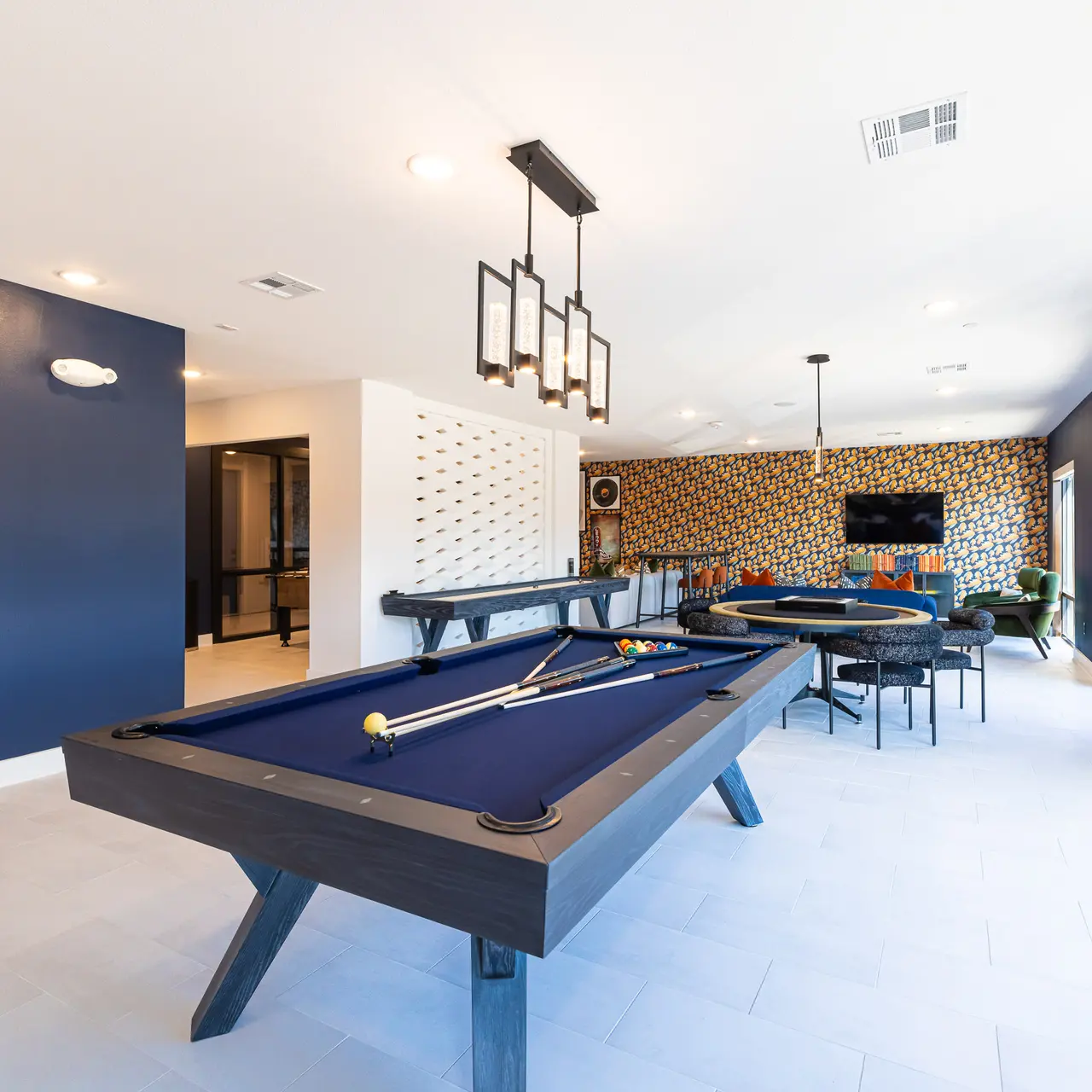 A spacious recreational room featuring a blue billiard table, modern lighting fixtures, and a stylish wallpapered wall. There are additional seating areas and a large window allowing natural light to fill the space.