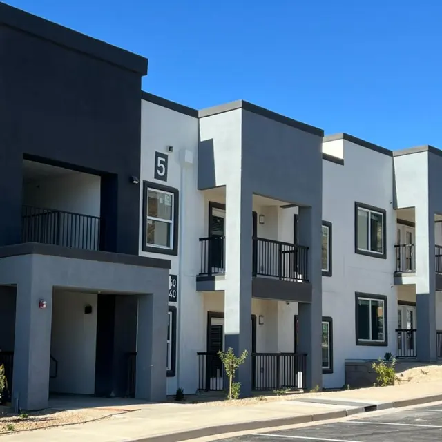 A modern apartment building with a mix of gray and white exterior walls, featuring multiple balconies and large windows under a clear blue sky.