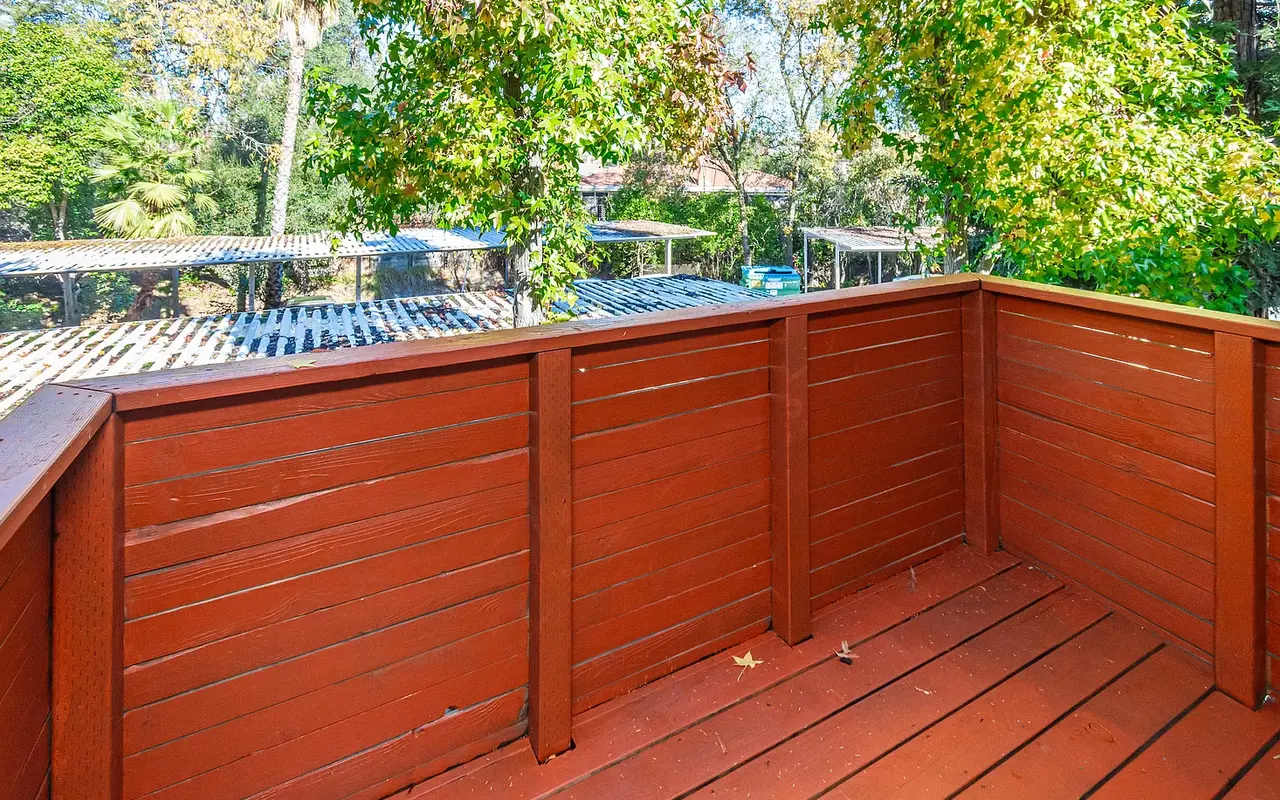 A wooden deck with reddish-brown flooring and railing, surrounded by greenery. Trees and other structures can be seen in the background.