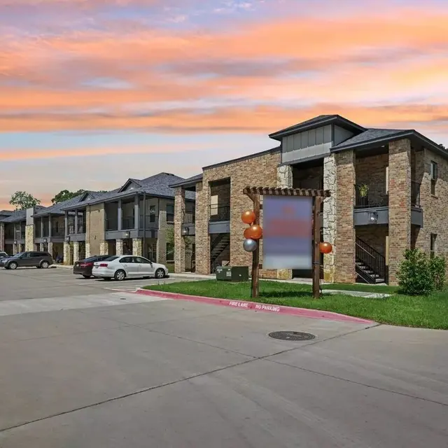 A view of a modern apartment complex featuring stone exteriors and multiple stories, with parked cars in the foreground and a vibrant sunset in the background.