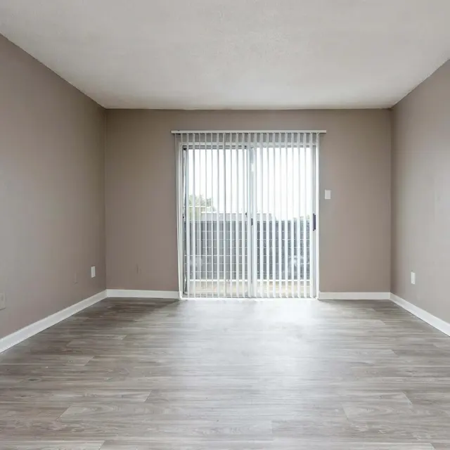 An empty room with light-colored walls and flooring, featuring a sliding glass door with vertical blinds.