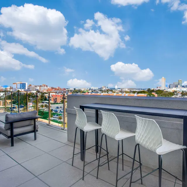 A modern rooftop lounge area featuring bar stools, a table, and seating, overlooking a city skyline with bright blue skies and fluffy clouds.