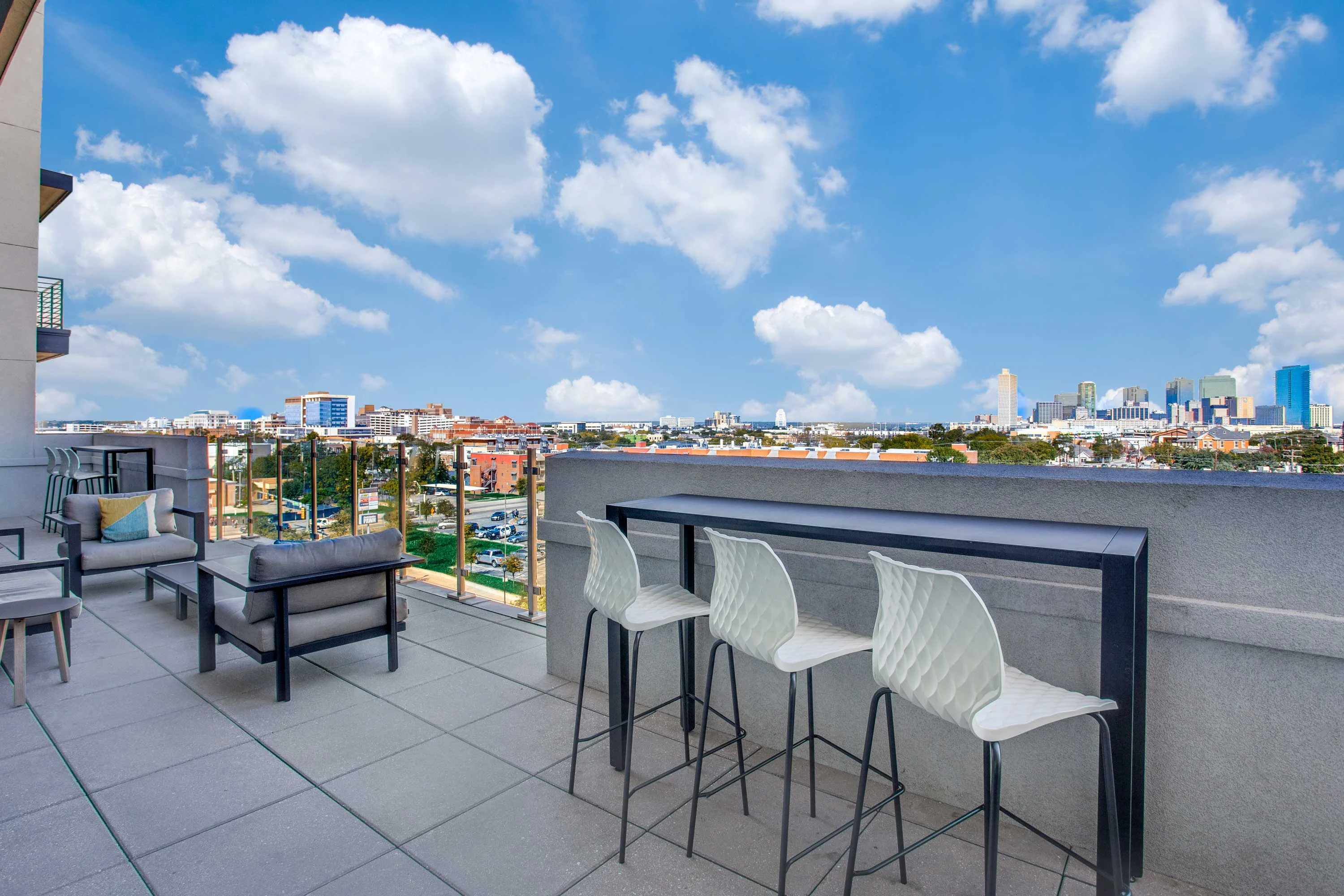 A modern rooftop lounge area featuring bar stools, a table, and seating, overlooking a city skyline with bright blue skies and fluffy clouds.