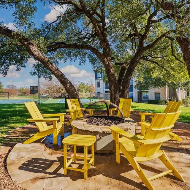 A cozy outdoor firepit area with yellow adirondack chairs arranged in a circle around a stone firepit. Lush greenery surrounds the space, with large trees providing shade and a grassy area nearby. A basketball court is visible in the background, along with a residential building.