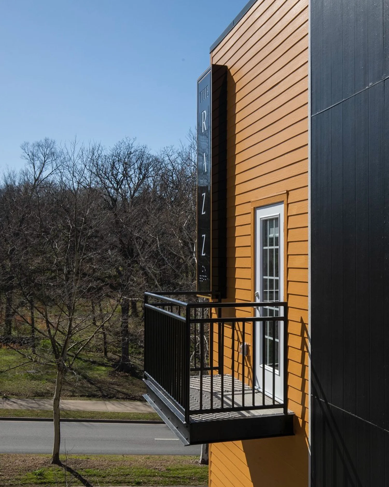 A close-up of a small balcony attached to an orange and black exterior building, overlooking a wooded area. The balcony features a railing and is bathed in natural light from a clear blue sky.