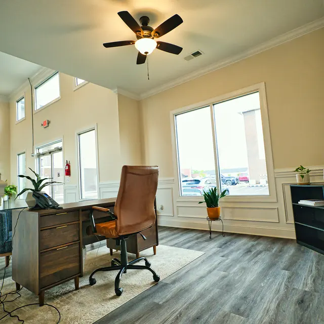 A modern office space featuring a wooden desk with a leather chair, a ceiling fan, and large windows letting in natural light.
