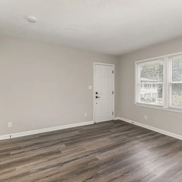 An empty living room with light gray walls and a wooden floor, featuring a door on the left and windows on the right that let in natural light.