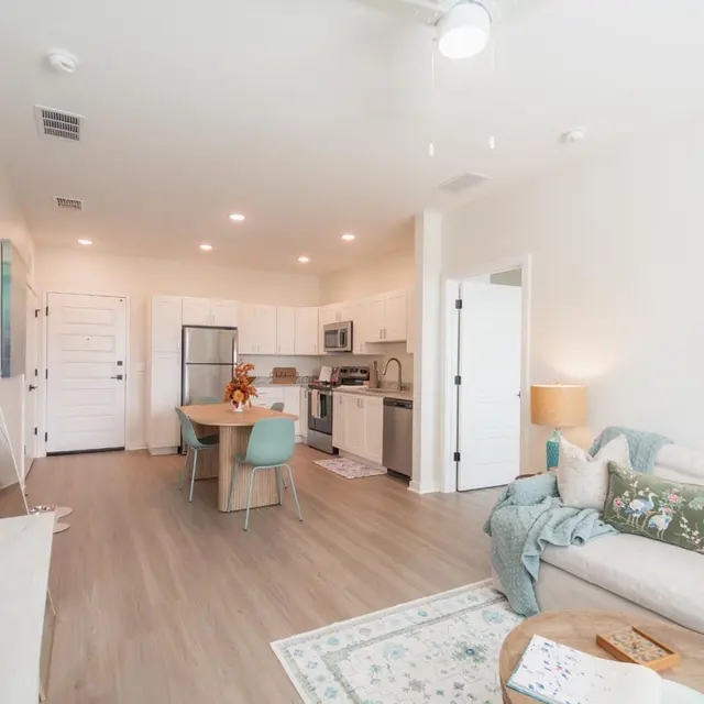 A bright, modern living room featuring a light-colored couch, a small dining table with chairs, and a well-equipped kitchen in the background. A colorful abstract art piece decorates the wall.