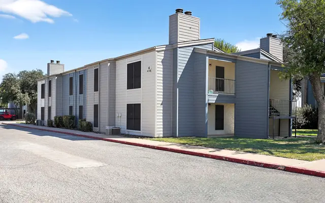 Exterior view of an apartment complex with a paved road in front, featuring multiple units and greenery.