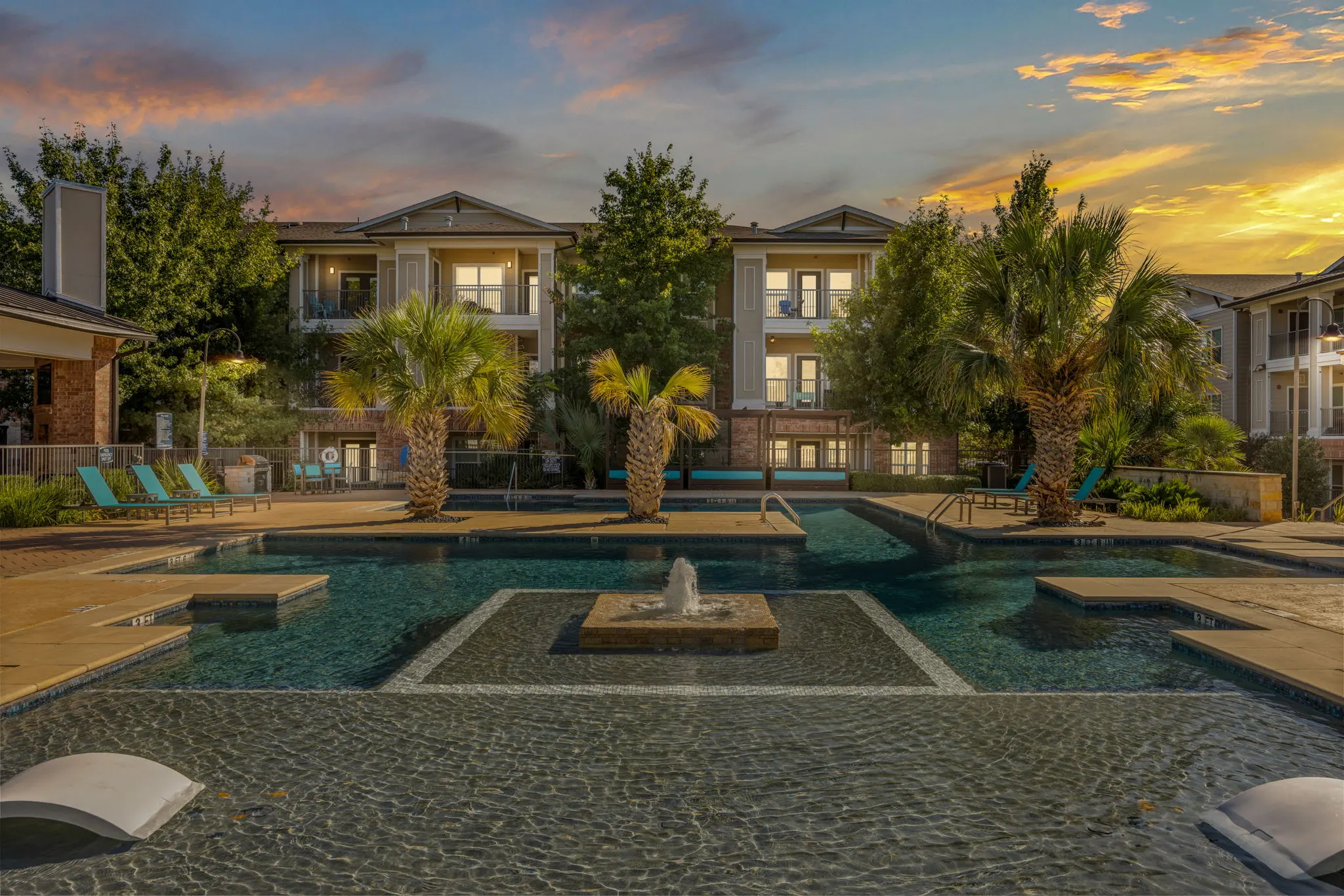 Charming Apartment Pool Area at Sunset A serene pool area in an apartment complex with palm trees, lounge chairs, and a fountain, set against a beautiful sunset sky.