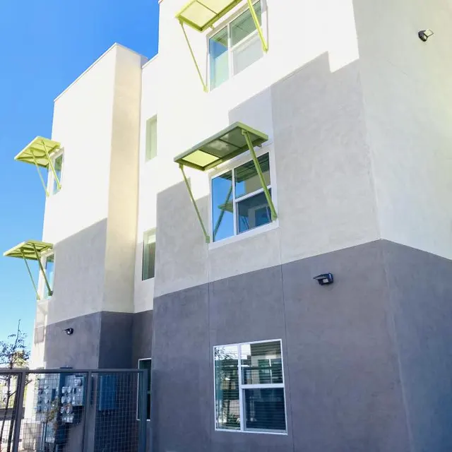 Contemporary Building Design A modern three-story building with a light-colored facade and unique green window canopies. The building is set against a clear blue sky and features a fenced area at the side.
