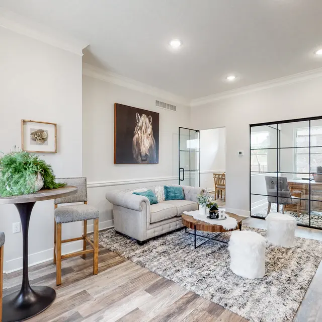 A modern living room with a comfortable sofa, coffee table, and bar stools. There is a decorative plant and framed art on the walls. A glass partition separates a workspace in the background.