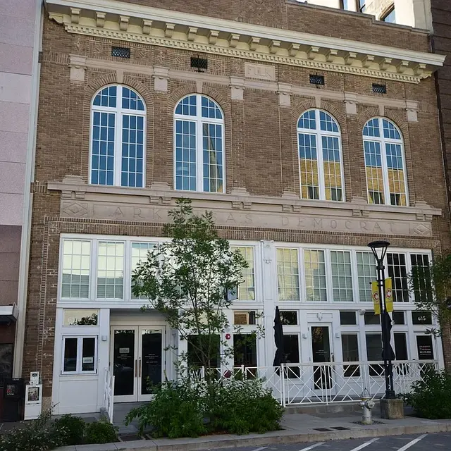 A historical brick building with large windows and ornamental detailing, featuring a white entrance with plants around it.