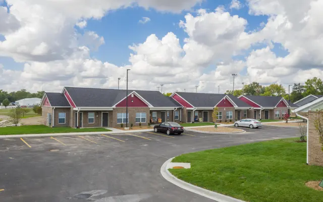 A view of a modern single-story housing complex with several units and parked cars situated in a green area under a partly cloudy sky.