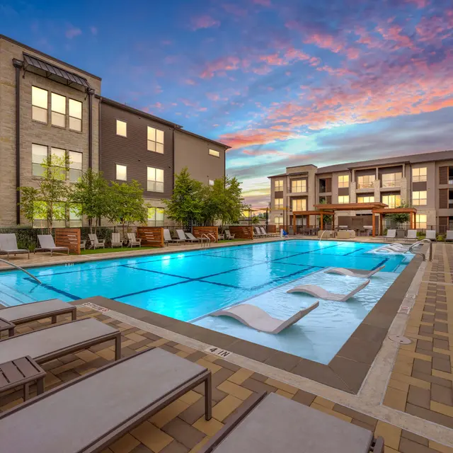 A modern apartment complex with a swimming pool at sunset. The pool is surrounded by lounge chairs, and the sky displays a colorful sunset with shades of orange and blue.