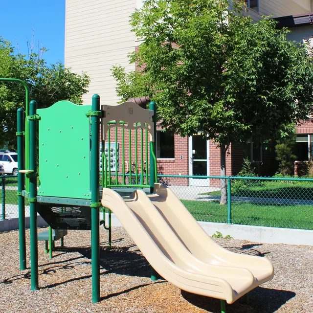 A playground structure featuring two beige slides, surrounded by gravel and greenery.