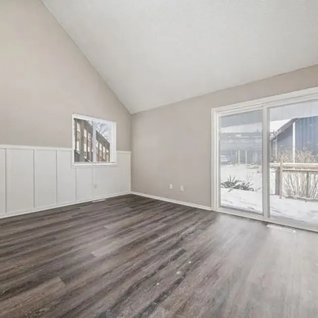 A spacious, empty room with wooden flooring and high vaulted ceilings. Natural light streams in through a large sliding door and a smaller window, illuminating the beige walls. The room has a simple, clean design with no furniture present.