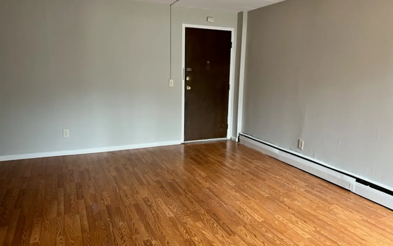 An empty room with wooden flooring, a light gray wall, and a closed brown door.