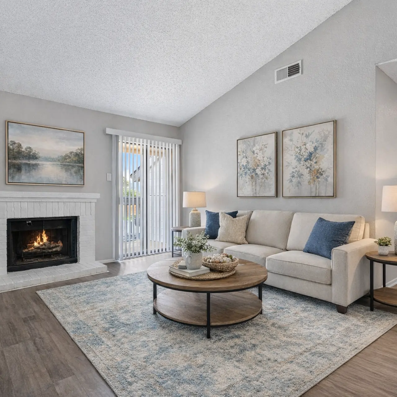 A modern living room featuring a light-colored sofa with blue and white pillows, a round coffee table, and a fireplace. The room has an airy feel with large windows and neutral walls. There are decorative pieces and artwork on the walls.