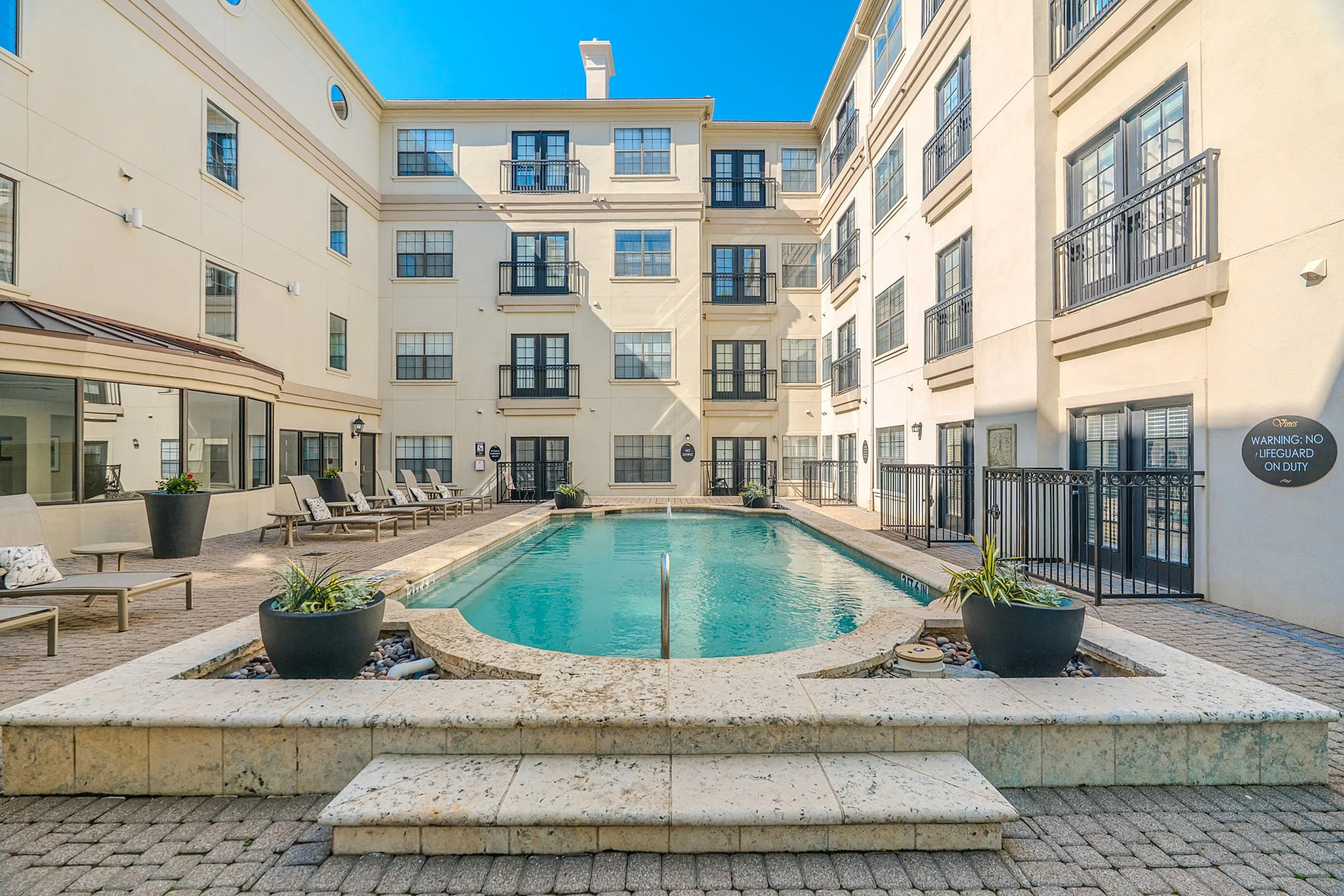 Apartment Complex Courtyard with Pool A view of an apartment complex courtyard featuring a swimming pool, surrounded by potted plants and lounge chairs under a clear blue sky.
