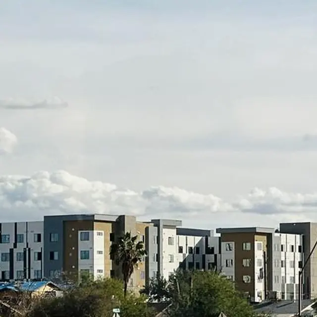 A modern apartment building with a light and dark color scheme, surrounded by green foliage and smaller houses in the foreground under a partly cloudy sky.