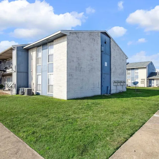 Exterior view of an apartment complex with two buildings, grassy areas, and a sidewalk under a blue sky with clouds.
