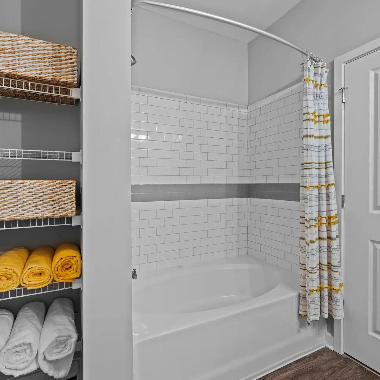 A modern bathroom featuring a bathtub with shower, white tile walls, and a door. There are neatly stacked yellow and white towels and decorative baskets on shelves.