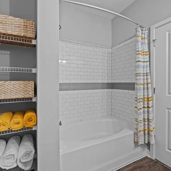 A modern bathroom featuring a bathtub with shower, white tile walls, and a door. There are neatly stacked yellow and white towels and decorative baskets on shelves.