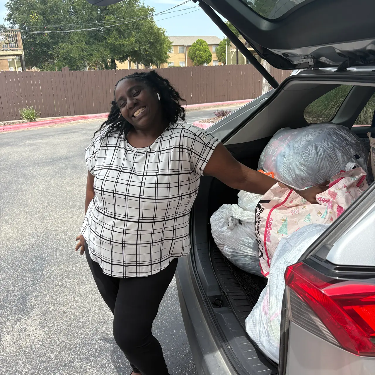 A woman poses next to the open trunk of a car filled with bags. She wears a plaid shirt and black pants, smiling at the camera.