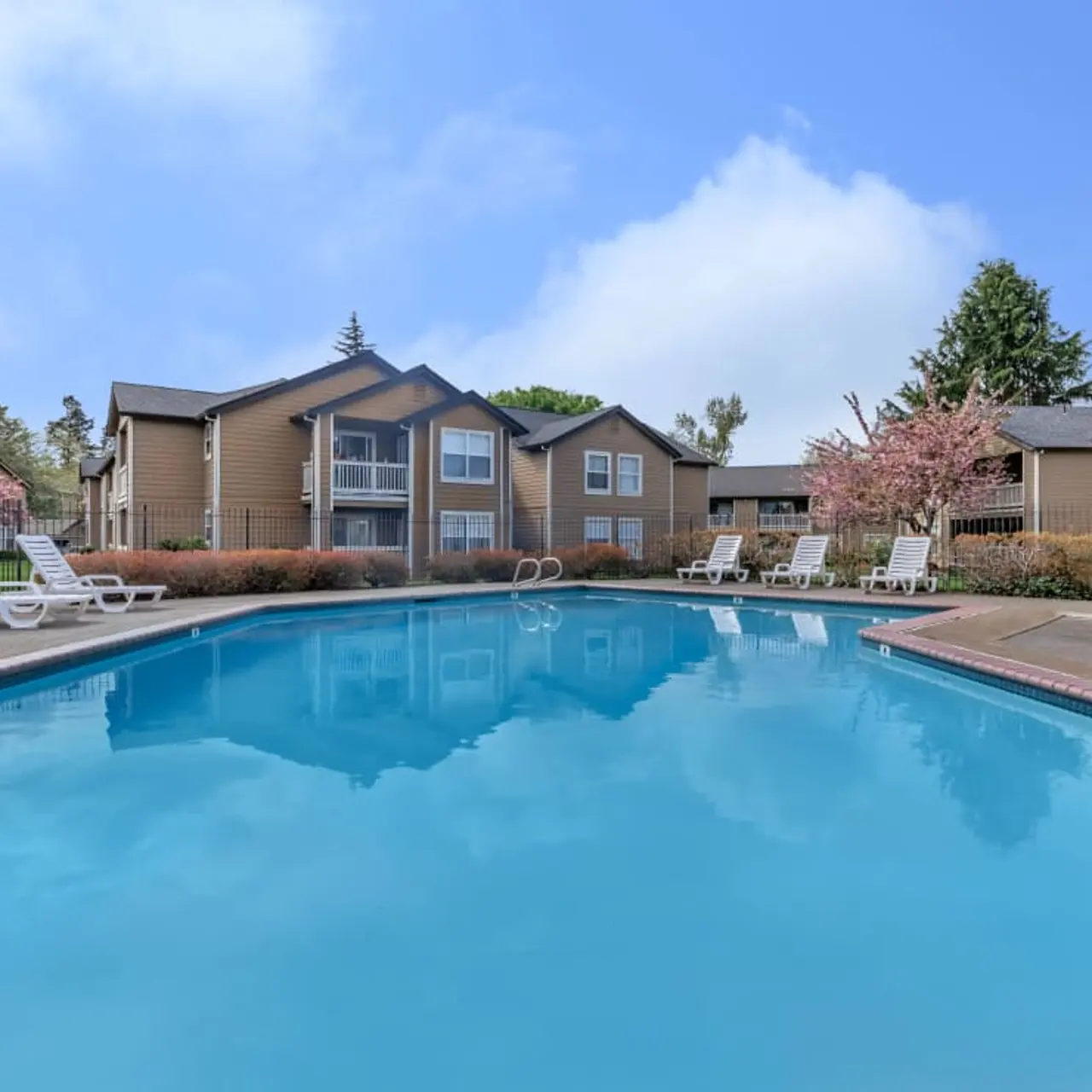 A residential pool area with clear blue water, surrounded by lounge chairs and shrubbery. Behind the pool are two-story apartment buildings with balconies, and blooming trees add a touch of nature to the scene under a clear blue sky.