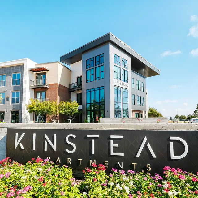 Modern apartment building with a sign reading 'Kinstead Apartments' in front, surrounded by colorful flowers and green landscaping.