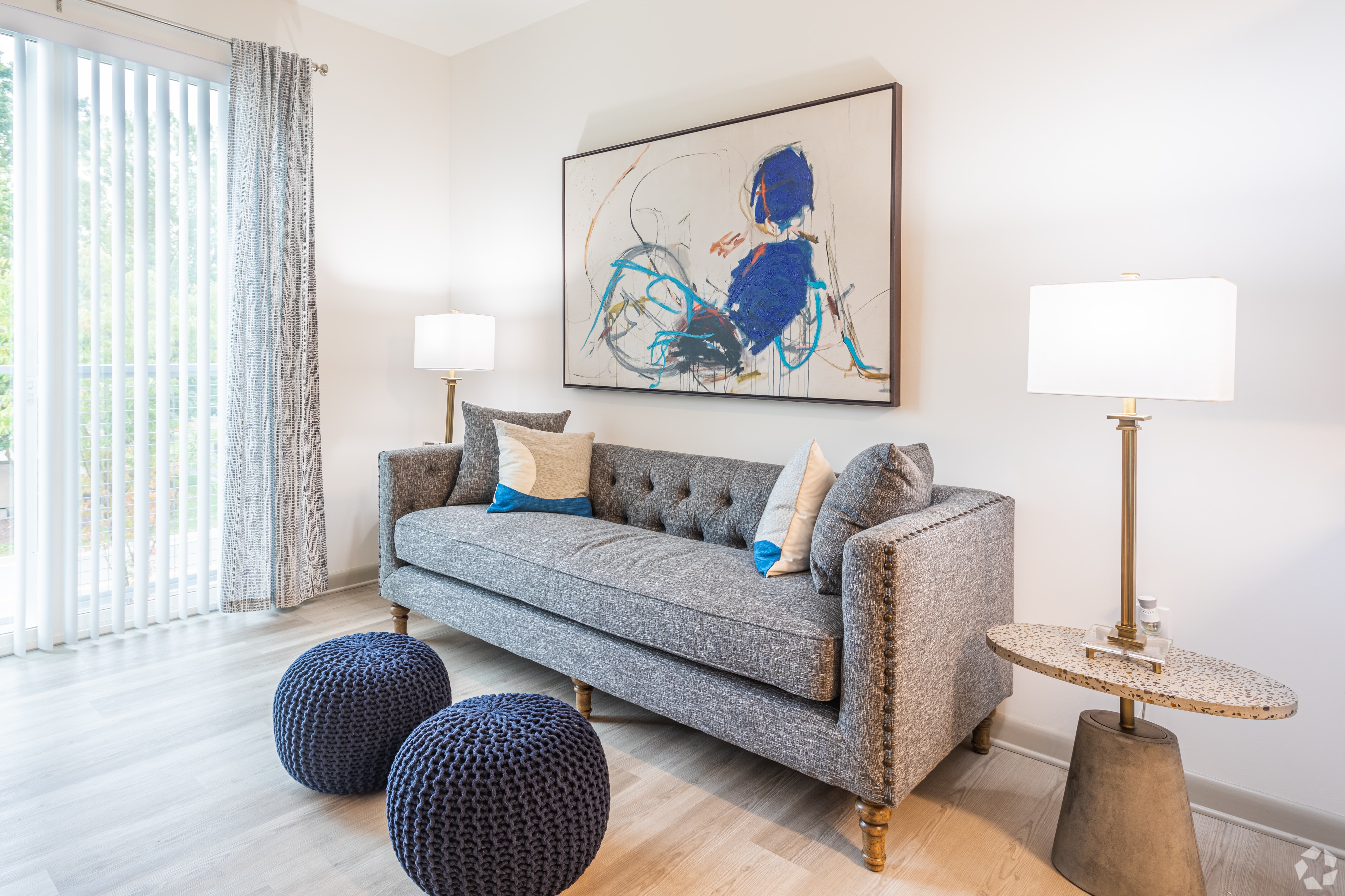 A modern living room featuring a gray tufted sofa with decorative pillows, two blue poufs on the floor, and a side table with a lamp. Large curtains cover a glass door leading outside, and an abstract painting hangs on the wall above the sofa.