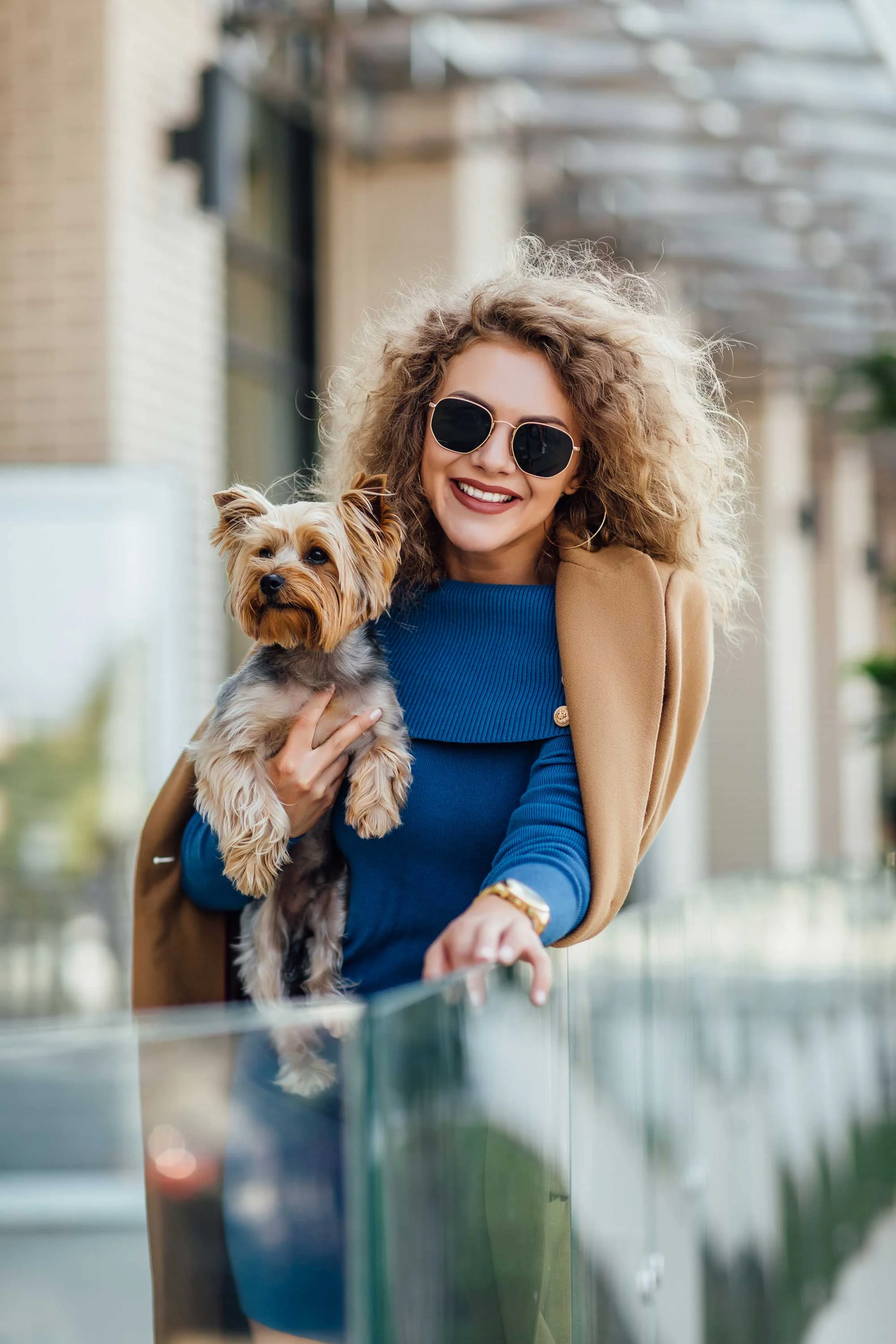 Stylish Woman with Dog A smiling woman with curly hair wearing sunglasses and holding a small dog while standing by a glass railing.