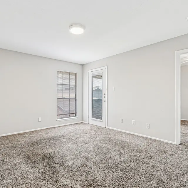 A spacious living room with light gray walls and plush carpeting, featuring a large window and a door leading outside. The room is well-lit, and there is an adjacent room visible with a ceiling fan.