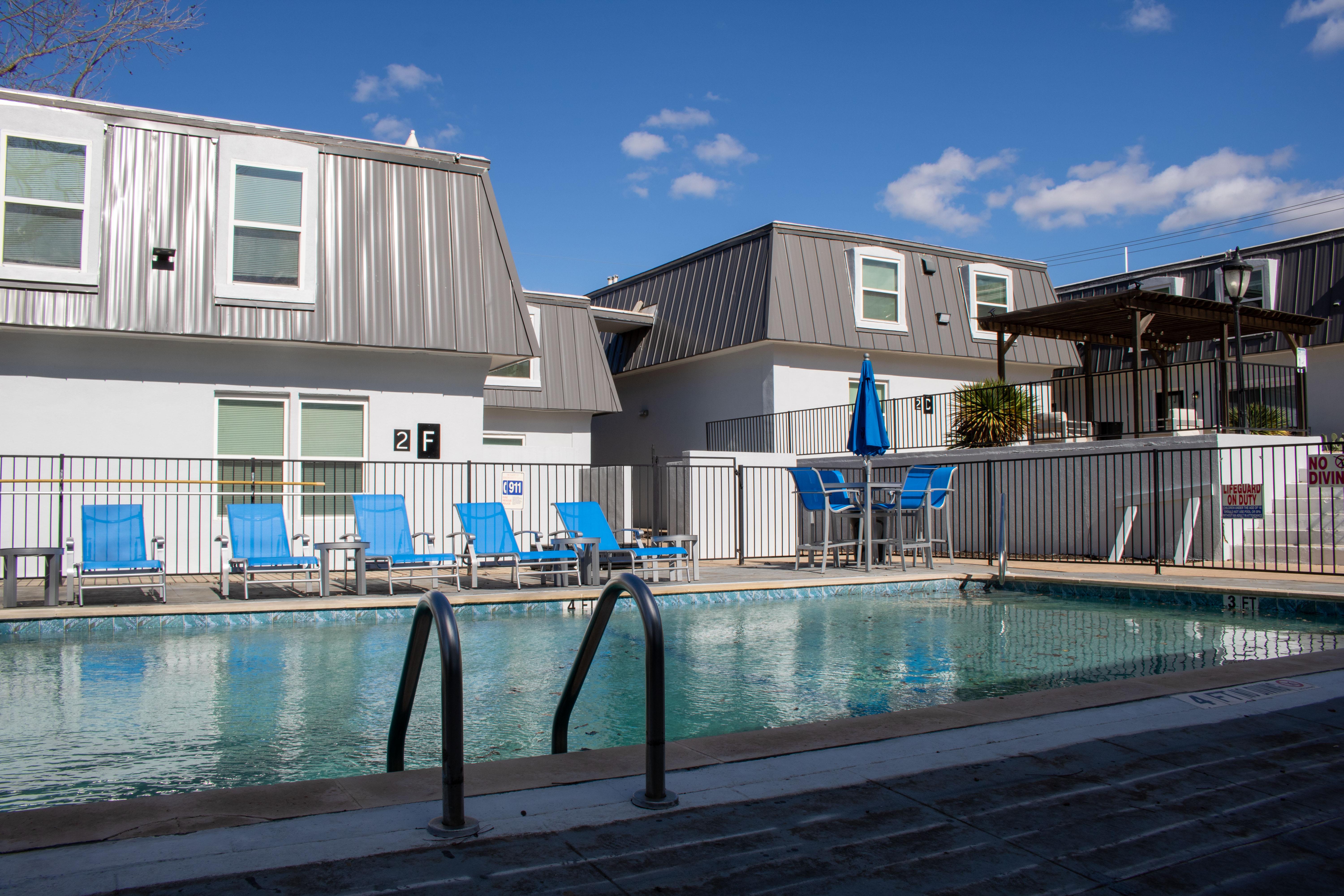 A swimming pool surrounded by blue chaise lounges and a fenced area. In the background, there are modern buildings with metal roofs and a shaded seating area. The sky is clear with a few clouds.