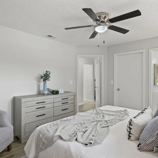 A modern bedroom featuring a queen-sized bed with decorative pillows and a light blanket, a gray couch with blue pillows, a nightstand, and a dresser. There are doors leading to adjacent rooms, and a ceiling fan is installed.
