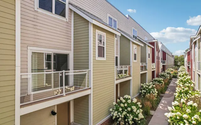 A modern apartment complex featuring a row of multi-colored buildings with balconies, surrounded by blooming flowers in a landscaped pathway.