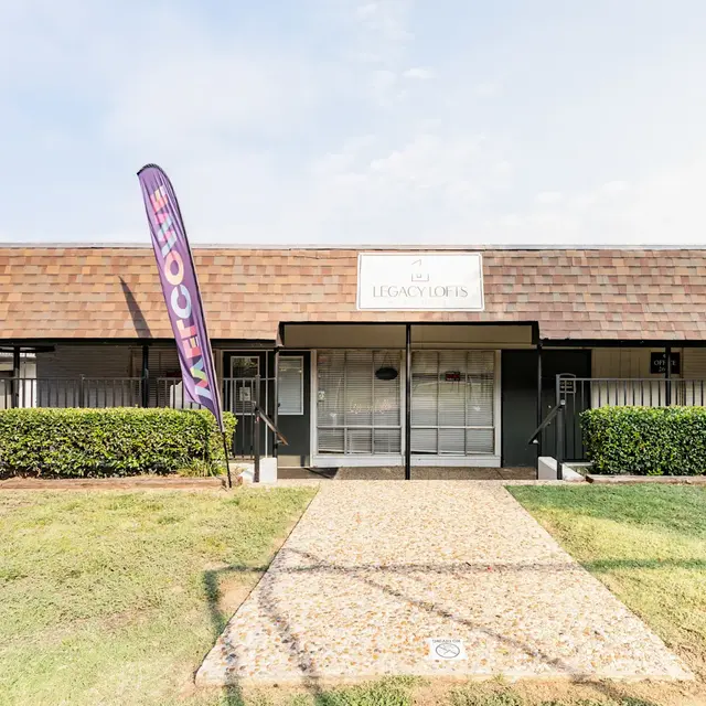 Front view of a building with a brown shingle roof and a sign that reads 'Legacy Works.' There are green bushes and a pebbled pathway leading to the entrance.