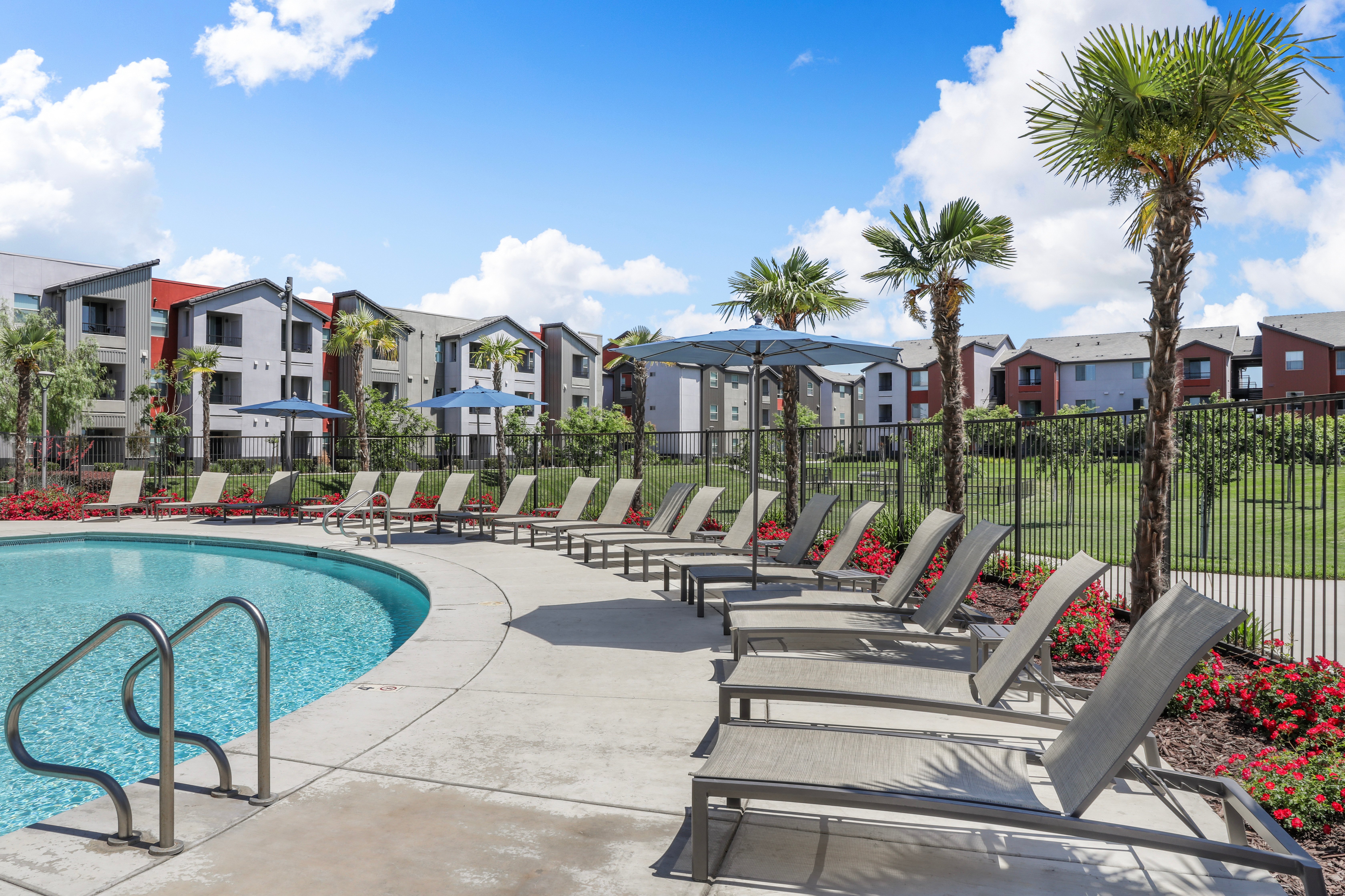 Relaxing Pool Area A sunny pool area with lounge chairs and a fenced yard, surrounded by palm trees and residential buildings in the background.