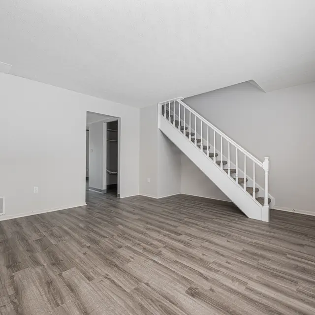 A spacious empty living room with light gray walls and brown wooden flooring. A staircase is visible on the right, leading to an upper level. There are two doorways to adjacent rooms, creating a sense of openness.