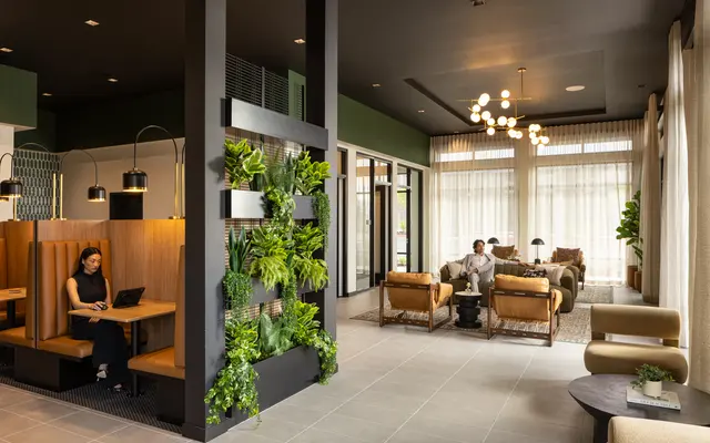Chic Modern Lounge Space A stylish lounge area featuring modern seating, greenery, and large windows allowing natural light in. One person is seated at a booth working on a laptop, while another couple is relaxing on a sofa in the background.